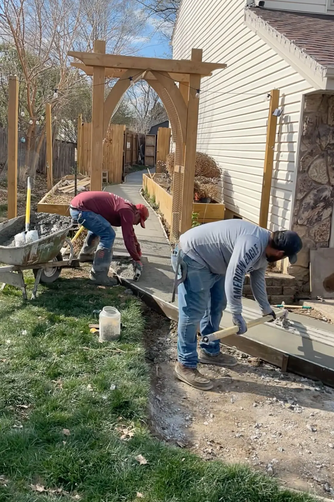 Two men working on pouring and smoothing fresh concrete for a curved garden path beneath a wooden arbor as part of an exterior house remodel. The path leads through a backyard garden space, bordered by raised beds and string lights overhead.